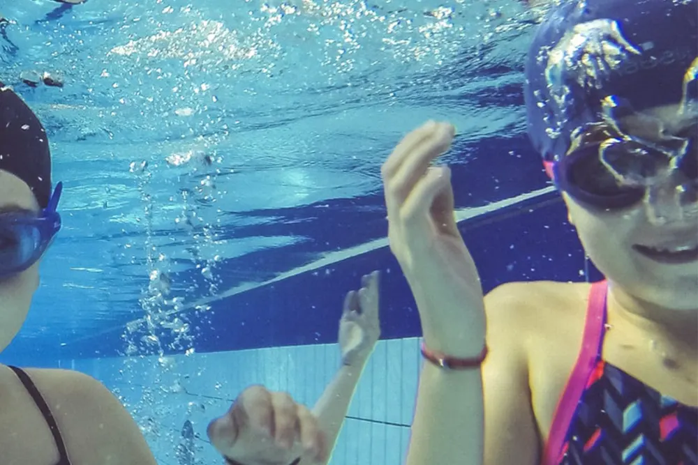 A group of smiling children underwater, wearing goggles and swim caps.
